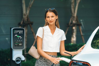 Genç bir kadın yazın yeşil sürdürülebilir şehir bahçesinde EV elektrikli arabasıyla seyahat ediyor. Kentsel sürdürülebilirlik yaşam tarzı yeşil temiz yeniden şarj edilebilir enerji elektrikli BEV araç iç organları