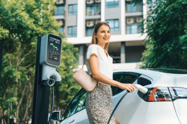 Genç bir kadın yazın yeşil sürdürülebilir şehir bahçesinde EV elektrikli arabasıyla seyahat ediyor. Kentsel sürdürülebilirlik yaşam tarzı yeşil temiz yeniden şarj edilebilir enerji elektrikli BEV araç iç organları