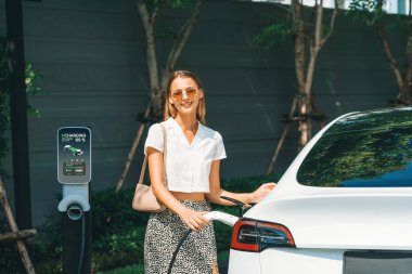 Genç bir kadın yazın yeşil sürdürülebilir şehir bahçesinde EV elektrikli arabasıyla seyahat ediyor. Kentsel sürdürülebilirlik yaşam tarzı yeşil temiz yeniden şarj edilebilir enerji elektrikli BEV araç iç organları