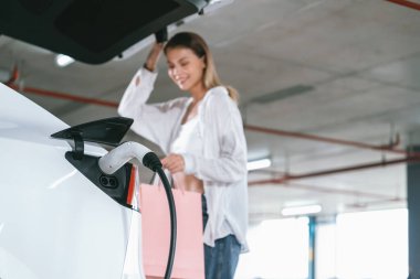 Genç bir kadın EV elektrikli arabasıyla alışveriş merkezinin otoparkına gidiyor şehir merkezinde elektrik iç organlarının yeşil temiz şarj edilebilir enerji ile sürdürülebilirlik yaşam tarzını gösteriyor.