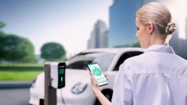 İş kadını, çevre yeşil şehir parkındaki şarj istasyonundan şarj olurken akıllı telefon ekranından EV batarya durum mobil uygulamasını kontrol ediyor. Elektrikli arabanın gelecekteki enerji sürdürülebilirliği.