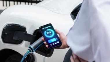 Akıllı telefon akü durumu arayüzünü elinde tutan iş kadını akıllı EV mobil uygulama ile otomobil parkındaki şarj istasyonundan elektrik şarj eden çevre dostu EV araba. İnceleyin