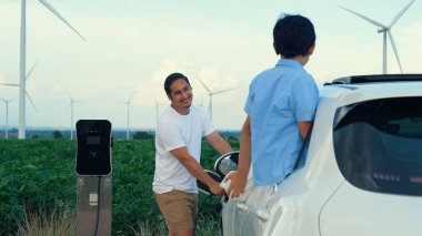 İlerici mutlu aile kavramı elektrikli araçla rüzgar çiftliğinde eğleniyor. Rüzgar türbini jeneratöründen gelen temiz yenilenebilir enerji ile şarj edilen elektrikli araç..