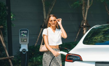 Genç bir kadın yazın yeşil sürdürülebilir şehir bahçesinde EV elektrikli arabasıyla seyahat ediyor. Kentsel sürdürülebilirlik yaşam tarzı yeşil temiz yeniden şarj edilebilir enerji elektrikli BEV araç iç organları