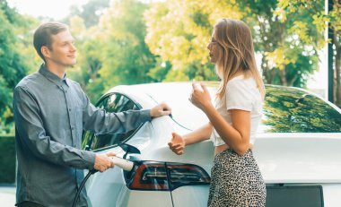 Genç çift yazın yeşil sürdürülebilir şehir bahçesinde EV elektrikli araba ile seyahat ederken, yeşil temiz yeniden şarj edilebilir elektrikli araç iç organları ile kentsel sürdürülebilirlik yaşam tarzını gösteriyor.