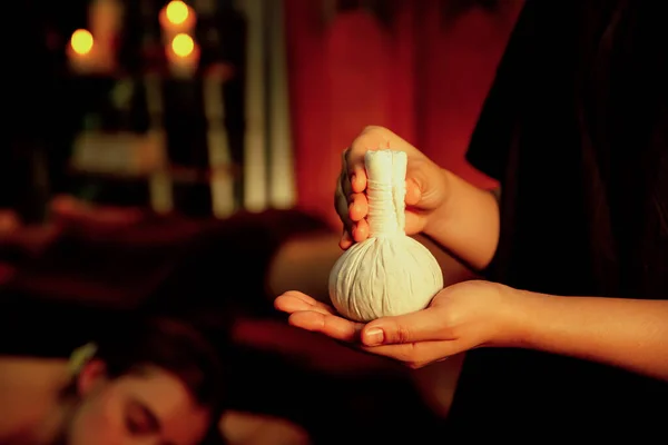 Hot herbal ball spa massage body treatment with masseurs hand hold or show herb bag at spa. Tranquil and serenity of aromatherapy recreation in warm lighting of candles at spa salon. Quiescent