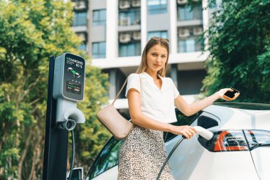 Genç bir kadın yazın yeşil sürdürülebilir şehir bahçesinde EV elektrikli arabasıyla seyahat ediyor. Kentsel sürdürülebilirlik yaşam tarzı yeşil temiz yeniden şarj edilebilir enerji elektrikli BEV araç iç organları