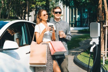 Genç çift yazın yeşil sürdürülebilir şehir bahçesinde EV elektrikli araba ile seyahat ederken, yeşil temiz yeniden şarj edilebilir elektrikli araç iç organları ile kentsel sürdürülebilirlik yaşam tarzını gösteriyor.