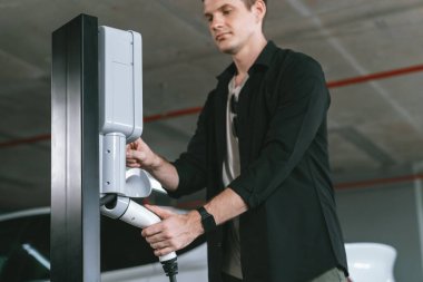 Genç adam EV elektrikli arabasıyla alışveriş merkezinin otoparkına gidiyor şehir merkezindeki şarj oluyor yeşil, temiz, yeniden şarj edilebilir elektrikli araç iç organları ile kentsel sürdürülebilirlik yaşam tarzını gösteriyor