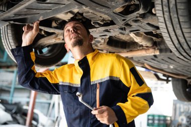 Araç tamircisi kaldırılmış aracın altından araba teftişi yapıyor. Üniformalı otomotiv teknisyeni araba akslarını ve şasi parçalarını dikkatle teşhis ediyor ve kontrol ediyor. Oxus.