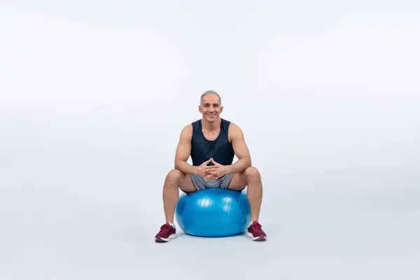 Full body length shot athletic and sporty senior man with fitness exercising ball in sitting posture on isolated background. Healthy active and body care lifestyle after retirement. Clout