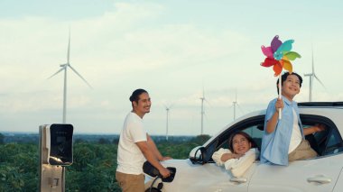 İlerici mutlu aile kavramı elektrikli araçla rüzgar çiftliğinde eğleniyor. Rüzgar türbini jeneratöründen gelen temiz yenilenebilir enerji ile şarj edilen elektrikli araç..