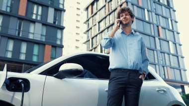 Takım elbiseli, hırslı bir işadamı elektrik kablosu ve yenilenebilir enerji enerjili bir elektrikli araçla şarj istasyonunda dururken elektrikli aracına yaslanıyor..
