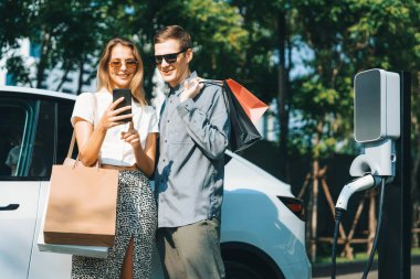 Genç çift yazın yeşil sürdürülebilir şehir bahçesinde EV elektrikli araba ile seyahat ederken, yeşil temiz yeniden şarj edilebilir elektrikli araç iç organları ile kentsel sürdürülebilirlik yaşam tarzını gösteriyor.