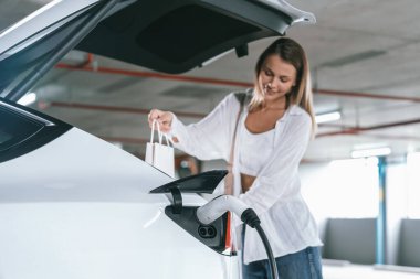 Genç bir kadın EV elektrikli arabasıyla alışveriş merkezinin otoparkına gidiyor şehir merkezinde elektrik iç organlarının yeşil temiz şarj edilebilir enerji ile sürdürülebilirlik yaşam tarzını gösteriyor.