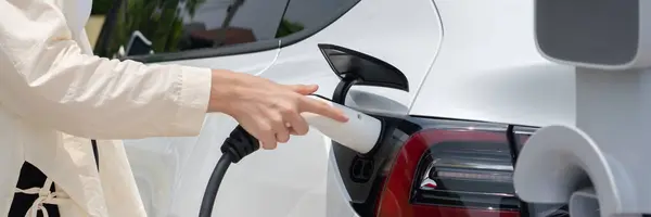 Young woman put EV charger to recharge electric cars battery from charging station. Alternative energy and rechargeable EV car for sustainable environmental friendly travel concept.Panorama Expedient