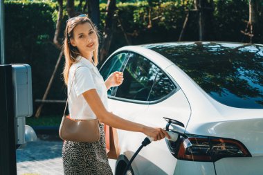 Genç bir kadın yazın yeşil sürdürülebilir şehir bahçesinde EV elektrikli arabasıyla seyahat ediyor. Kentsel sürdürülebilirlik yaşam tarzı yeşil temiz yeniden şarj edilebilir enerji elektrikli BEV araç iç organları