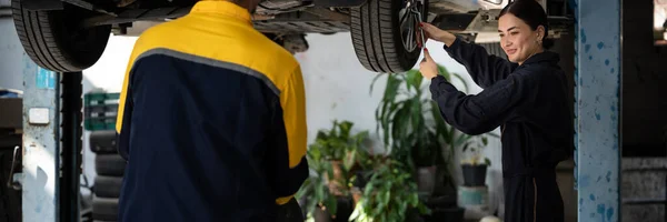 Vehicle mechanic conduct car inspection from beneath lifted vehicle. Automotive service technician in uniform carefully diagnosing and checking cars axles and undercarriage components. Panoram Oxus