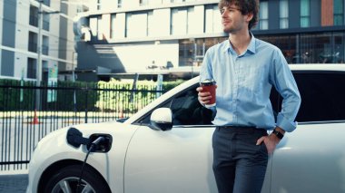 İlerici işadamı, şehir merkezinde dolaşmadan önce şarj cihazından elektrikli arabasına şarj prizi takıyor. Çevre dostu şarj edilebilir araba, sürdürülebilir ve temiz enerjiyle çalışıyor..