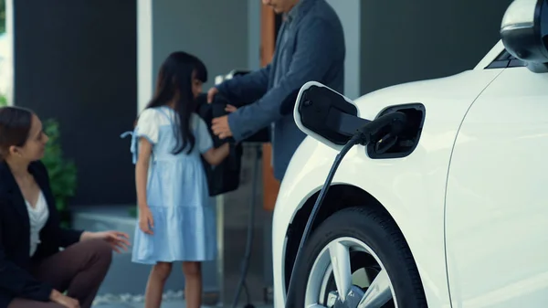 Progressive parents with electric vehicle and home charging station. Happy family with daughter giving each other high fives before leave for school. Alternative future transportation concept of EVs