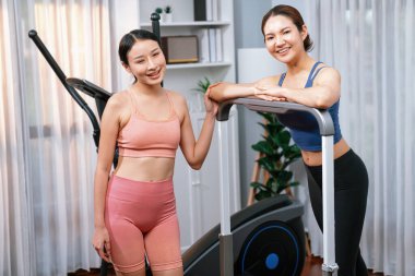Enerjik ve güçlü atletik Asyalı kadın evde spor arkadaşı ya da antrenörü ile birlikte eliptik koşu makinesiyle koşuyor. Formda fiziğin takibi ve sağlıklı yaşam tarzına bağlılık. Kuvvetli