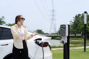 EV araba aküsünü şarj eden genç bir kadın elektrik şebekesine bağlı elektrik santralinde çevre dostu araç kullanımı için elektrik endüstrisi olarak telefonla konuşurken. Yararlı
