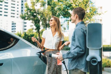 Genç çift yazın yeşil sürdürülebilir şehir bahçesinde EV elektrikli araba ile seyahat ederken, yeşil temiz yeniden şarj edilebilir elektrikli araç iç organları ile kentsel sürdürülebilirlik yaşam tarzını gösteriyor.
