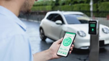 Akıllı telefon ekran bataryasını elinde tutan iş adamı akıllı EV mobil uygulama ile otomobil parkındaki şarj istasyonundan elektrik şarj eden çevre dostu EV otomobil. İnceleyin