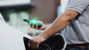 Modern iş adamı akıllı telefonun 3D hologramından batarya durumunu kontrol ederken EV 'si şarj olan enerjisini kontrol ediyor. Gelecekteki EV otomotiv teknolojisinin yenilikçi teknolojik gelişimi. İnceleyin