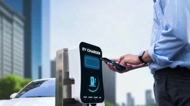 İşadamı, EV batarya durum hologramını akıllı telefon uygulamasından kontrol ederken EV aracını EV 'nin şarj ettiği çevre yeşil şehir parkından ağaçla şarj ediyor. Geleceğin yenilikçi enerji sürdürülebilirliği. İnceleyin