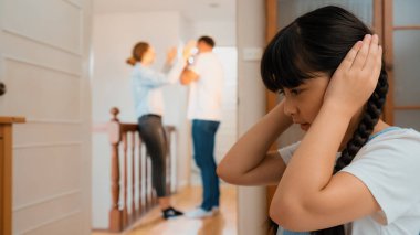 Stresli ve mutsuz genç kız köşeye kıvrılıp kulaklarını kapatarak arka planda ebeveyninin tartışmasını engelliyor. Evde aile içi şiddet ve travmatik çocukluk depresyona dönüşür. Eşzamanlama