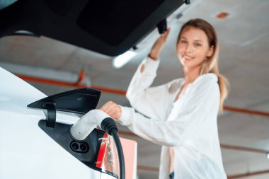 Genç bir kadın EV elektrikli arabasıyla alışveriş merkezinin otoparkına gidiyor şehir merkezinde elektrik iç organlarının yeşil temiz şarj edilebilir enerji ile sürdürülebilirlik yaşam tarzını gösteriyor.
