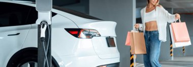 Genç bir kadın EV elektrikli arabasıyla alışveriş merkezinin otoparkına gidiyor şehir merkezinde elektrik iç organlarının yeşil temiz şarj edilebilir enerji ile sürdürülebilirlik yaşam tarzını gösteriyor.