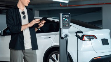 Genç adam EV elektrikli arabasıyla alışveriş merkezinin otoparkına gidiyor şehir merkezindeki şarj oluyor yeşil, temiz, yeniden şarj edilebilir elektrikli araç iç organları ile kentsel sürdürülebilirlik yaşam tarzını gösteriyor