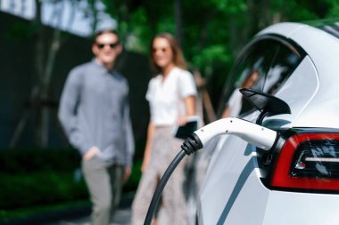 Genç çift yazın yeşil sürdürülebilir şehir bahçesinde EV elektrikli araba ile seyahat ederken, yeşil temiz yeniden şarj edilebilir elektrikli araç iç organları ile kentsel sürdürülebilirlik yaşam tarzını gösteriyor.