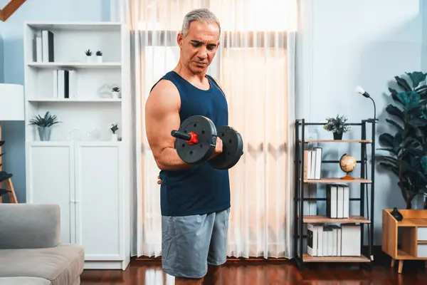 Athletic and sporty senior man engaging in body workout routine with lifting dumbbell at home as concept of healthy fit body with body weight lifestyle after retirement. Clout