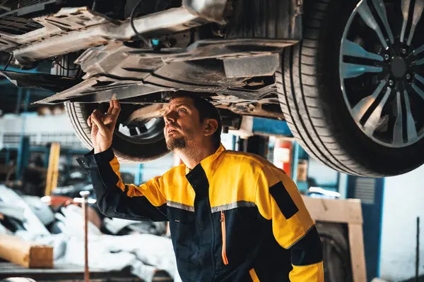 Vehicle mechanic conduct car inspection from beneath lifted vehicle. Automotive service technician in uniform carefully diagnosing and checking cars axles and undercarriage components. Oxus