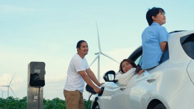 İlerici mutlu aile kavramı elektrikli araçla rüzgar çiftliğinde eğleniyor. Rüzgar türbini jeneratöründen gelen temiz yenilenebilir enerji ile şarj edilen elektrikli araç..