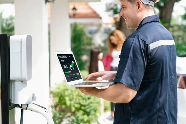 Qualified technician working on home EV charging station installation, making troubleshooting and configuration setup on charging system with laptop for EV at home with the family present. Synchronos