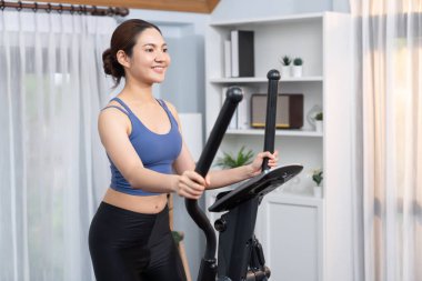 Enerjik ve güçlü atletik Asyalı kadın evde eliptik koşu makinesiyle koşuyor. Formda fiziğin takibi ve ev çalışması ve eğitimle sağlıklı yaşam tarzına bağlılık. Kuvvetli