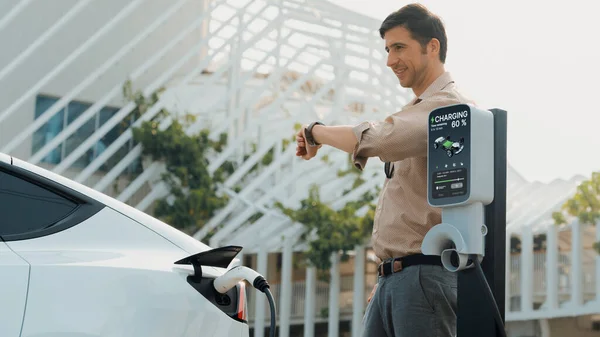 スマートウォッチで時間をチェックしている若い男は Ev充電器が市内モール駐車場の充電ステーションから充電する 持続可能なエコフレンドリーな都市旅行のための充電可能なEvカー エクスペディエント — ストック写真