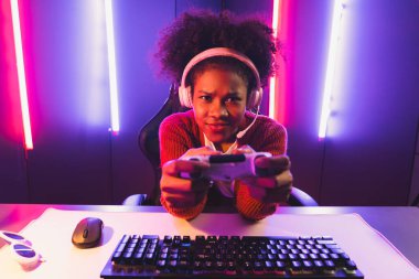 Oyun flamasının sunucu kanalı, joystick ile online oyun oynayan Afrikalı kız, mikrofondan izleyici medyasıyla konuşuyor. Esport yetenekli takım oyuncuları neon renkli aydınlatma odasında. Tatlandırıcı.