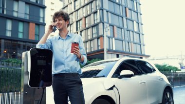 Takım elbiseli, hırslı bir işadamı elektrik kablosu ve yenilenebilir enerji enerjili bir elektrikli araçla şarj istasyonunda dururken elektrikli aracına yaslanıyor..