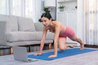 Spor kıyafetli Asyalı bir kadın antrenman rutini olarak spor minderinin üzerinde burpee yapıyor. Çekici kız sağlıklı yaşam tarzını online egzersiz videosuyla takip ediyor. Kuvvetli