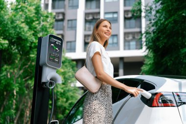 Genç bir kadın yazın yeşil sürdürülebilir şehir bahçesinde EV elektrikli arabasıyla seyahat ediyor. Kentsel sürdürülebilirlik yaşam tarzı yeşil temiz yeniden şarj edilebilir enerji elektrikli BEV araç iç organları