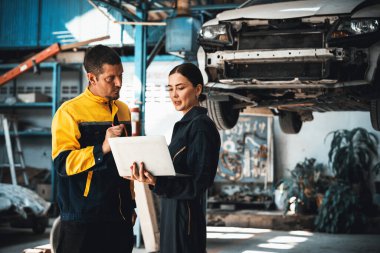 İki araç tamircisi birlikte çalışıyor, dizüstü bilgisayarla araba teftişi yapıyor. Üniformalı otomotiv servis teknisyeni hatayı tanımlamak için dikkatle sorun çıkartıyor. oxus