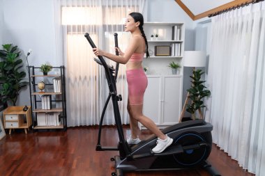 Enerjik ve güçlü atletik Asyalı kadın evde eliptik koşu makinesiyle koşuyor. Formda fiziğin takibi ve ev çalışması ve eğitimle sağlıklı yaşam tarzına bağlılık. Kuvvetli