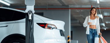 Genç bir kadın EV elektrikli arabasıyla alışveriş merkezinin otoparkına gidiyor şehir merkezinde elektrik iç organlarının yeşil temiz şarj edilebilir enerji ile sürdürülebilirlik yaşam tarzını gösteriyor.