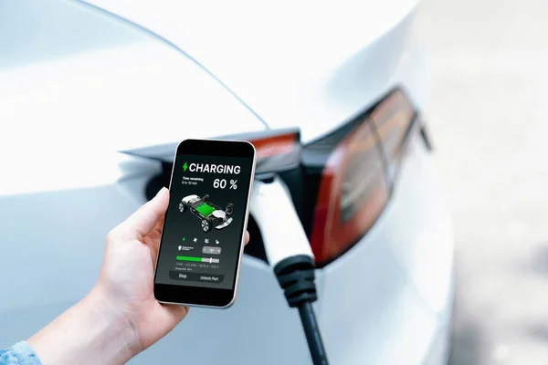 Hand insert EV charger plug into electric vehicle to recharge EV car, battery status display on smartphone EV application. Future alternative clean and sustainable energy for transportation. Perpetual