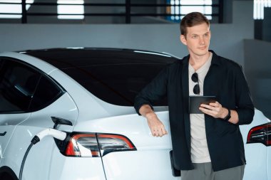 Genç adam EV elektrikli arabasıyla alışveriş merkezinin otoparkına gidiyor şehir merkezindeki şarj oluyor yeşil, temiz, yeniden şarj edilebilir elektrikli araç iç organları ile kentsel sürdürülebilirlik yaşam tarzını gösteriyor
